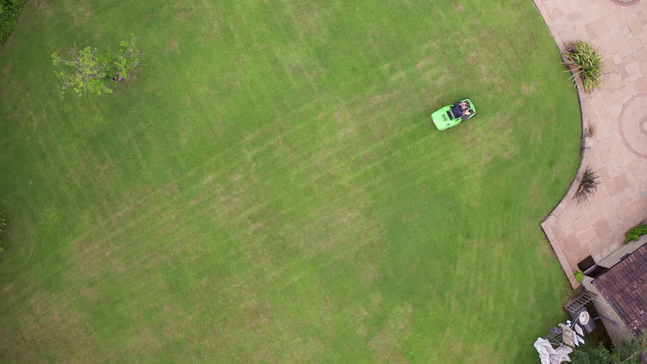Rising Aerial Shot of Man Mowing Lawn of Home on Summer’s Day using Ride On Lawnmower with Birds Eye View Perspective