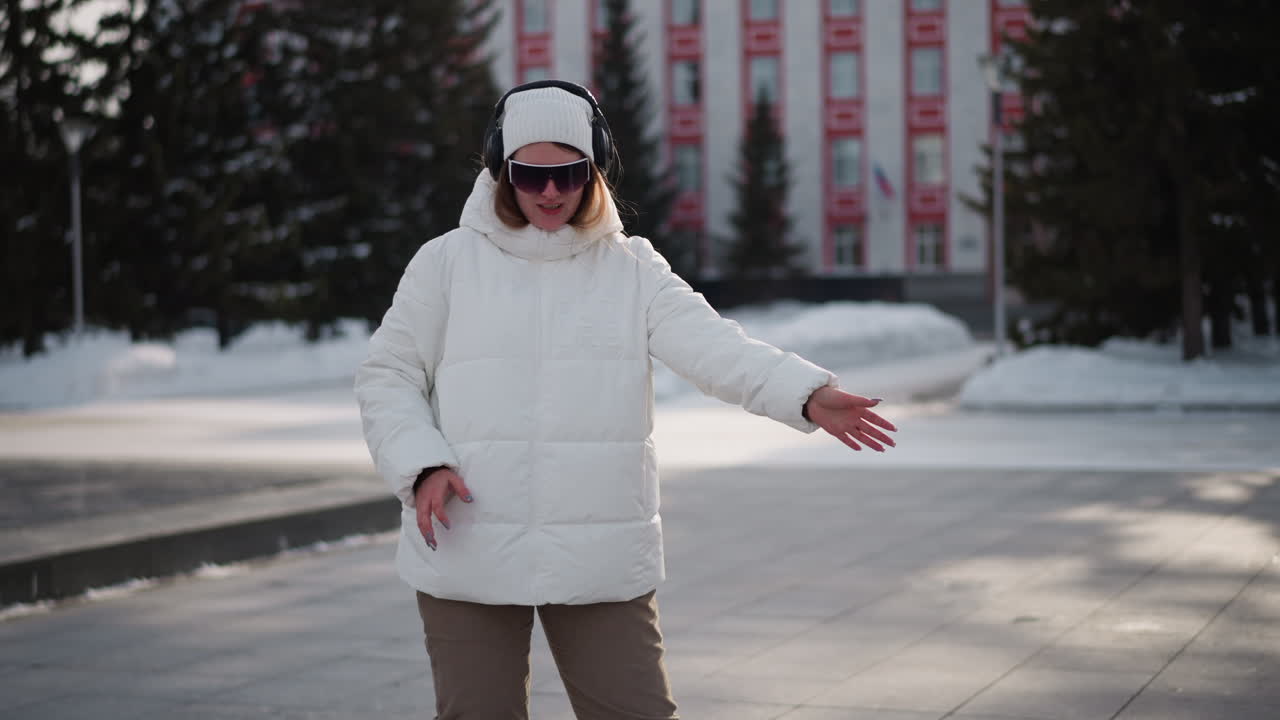 Teenager wearing white winter coat, beanie and headphones performs professional dance moves on snowy tiled plaza with urban backdrop under soft daylight quickly while pedestrians pass by