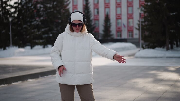 Teenager wearing white winter coat, beanie and headphones performs professional dance moves on snowy tiled plaza with urban backdrop under soft daylight quickly while pedestrians pass by