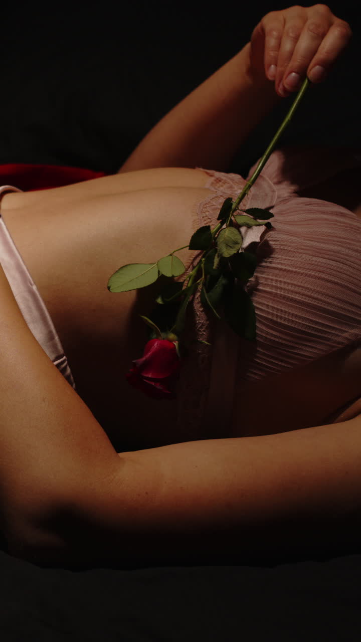 Bellissimo primo piano verticale di una donna attraente che accarezza il suo corpo con una rosa mentre indossa lingerie seducente a letto