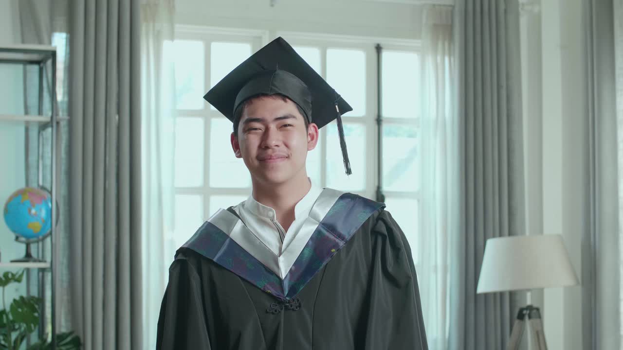 Asian Man Wearing A Graduation Gown And Cap Smiling To Camera In Living Room