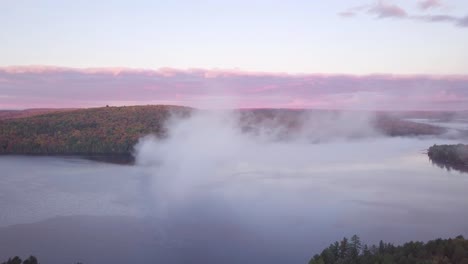 aerial sunrise wide shot flying through cloud fog showing misty lake and pink clouds and fall forest colors in kawarthas ontario canada