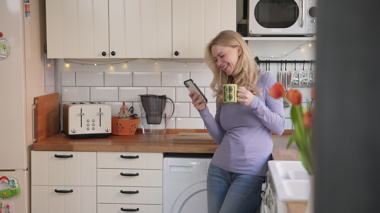 Woman in kitchen using smartphone and drinking from a cup