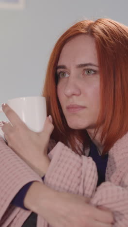 focused woman with ginger hair watches exciting tv show covering with pink blanket. housewife enjoys television program after work drinking hot tea closeup