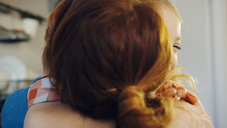 Close-up-of-the-cute-blonde-girl-hugging-her-mom-tight-and-smiling.-Rear-of-the-mother.-Portrait-shot.-Indoors