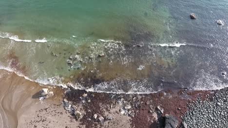 Aerial-overhead-descending-view-of-green-sea-and-white-foaming-waves-gently-breaking-on-the-rocky-sandy-shore-in-summer
