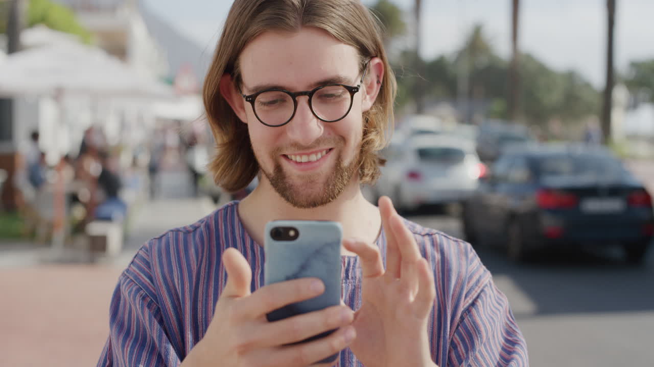 ritratto di un bel giovane che utilizza uno smartphone e si diverte a navigare online, inviare messaggi di testo, condividere vacanze sui social media, sullo sfondo di una vivace spiaggia urbana, indossando occhiali