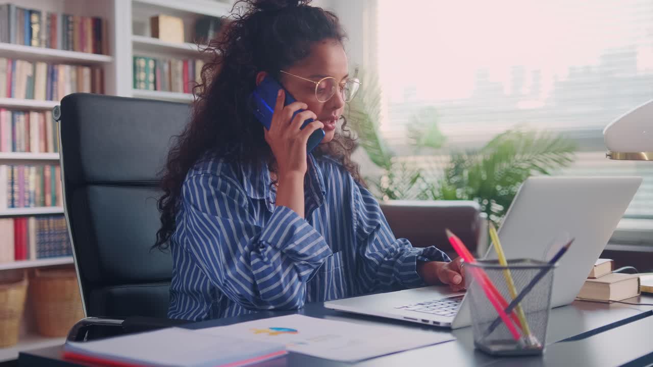 Ung indisk forretningskvinde taler i mobiltelefon og kigger på sin bærbare computer.