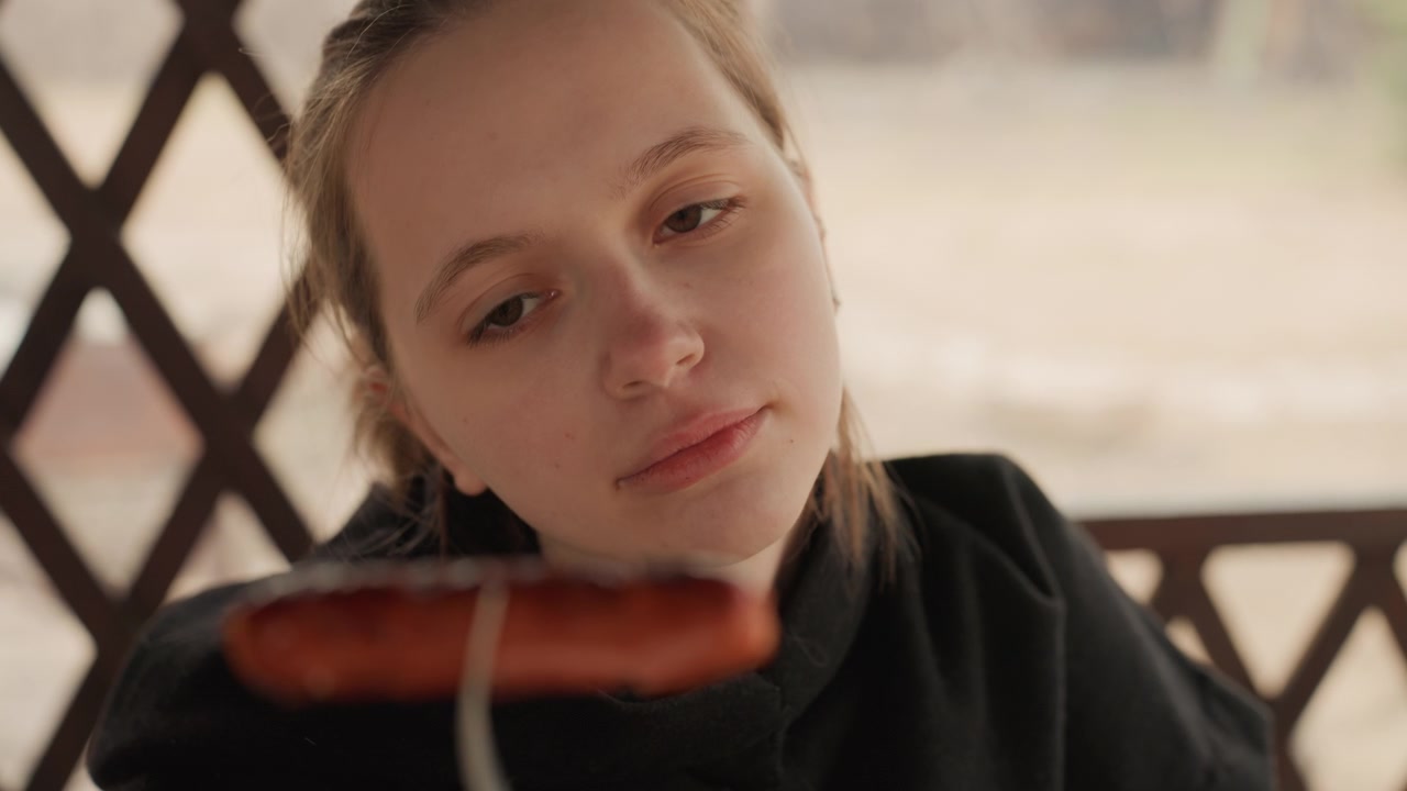 Giovane donna in abiti scuri tiene una salsiccia grigliata su una forchetta vicino al viso, sguardo abbassato in una pausa pensierosa prima di mangiare, con luce laterale calda, sfocatura dello sfondo morbida, ritratto intimo all'aperto.