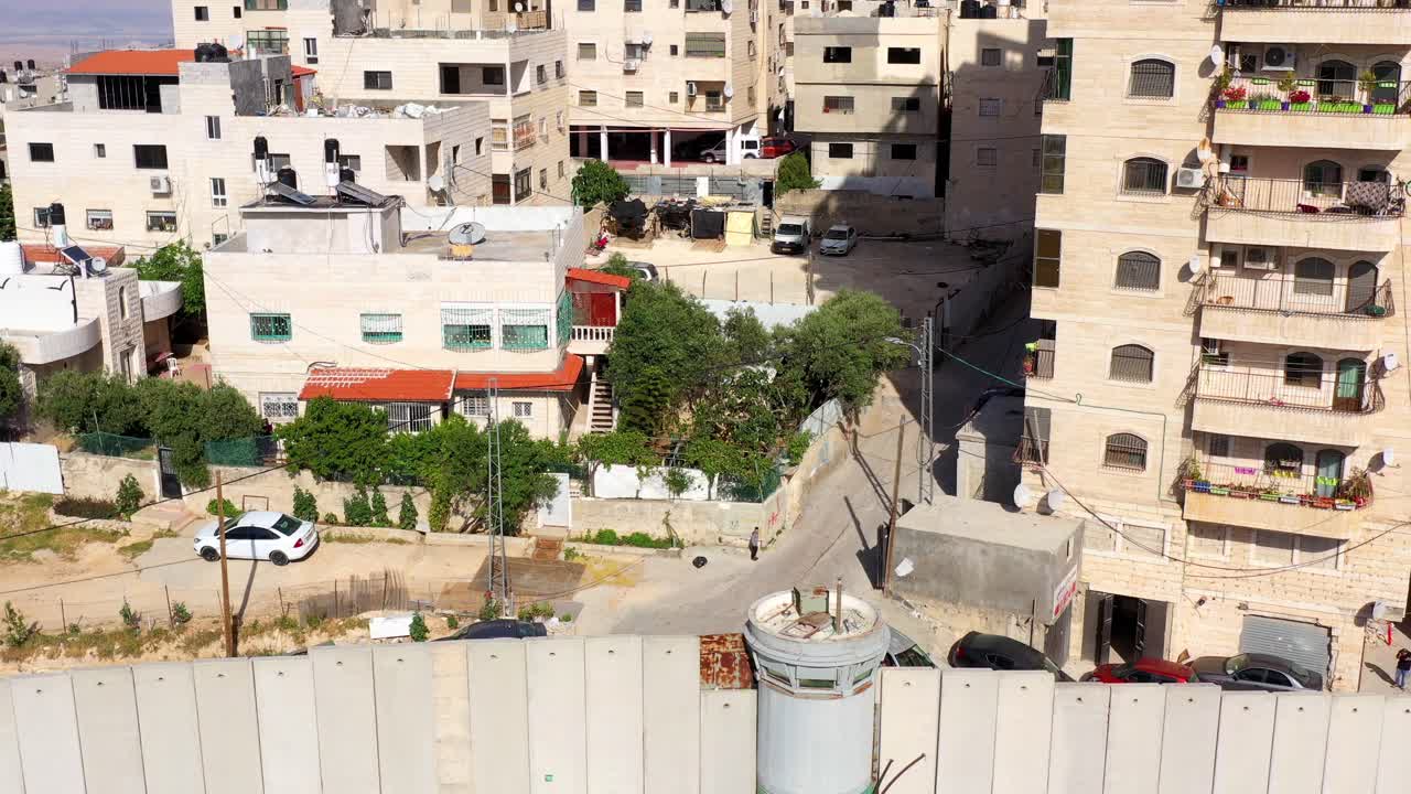 Aerial view of the Israeli Separation Barrier with a watchtower, flanked by residential buildings in a Middle Eastern urban landscape