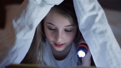 close-up view of cute little girl using a flashlight and reading a book with surprised expression under the blanket at night