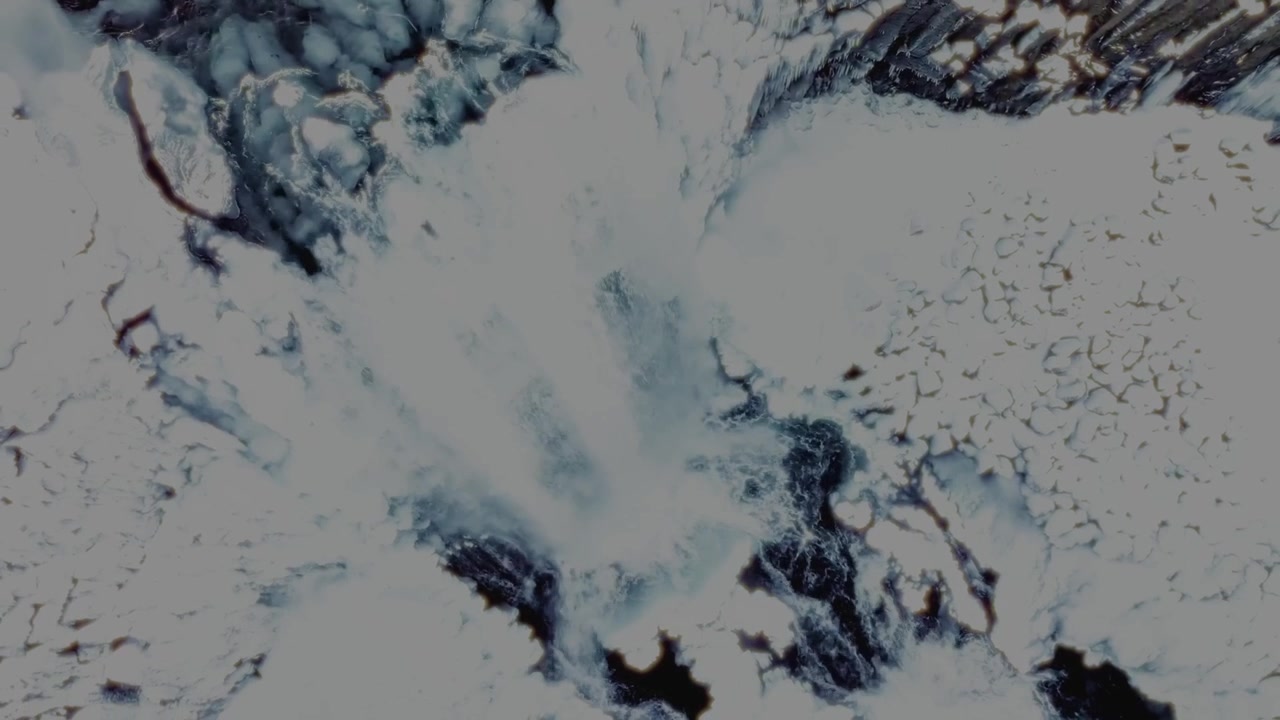 A breathtaking top-down drone shot of a partially frozen Icelandic waterfall surrounded by snow and ice-covered terrain. The bird's-eye perspective reveals the powerful flow of water cutting through t
