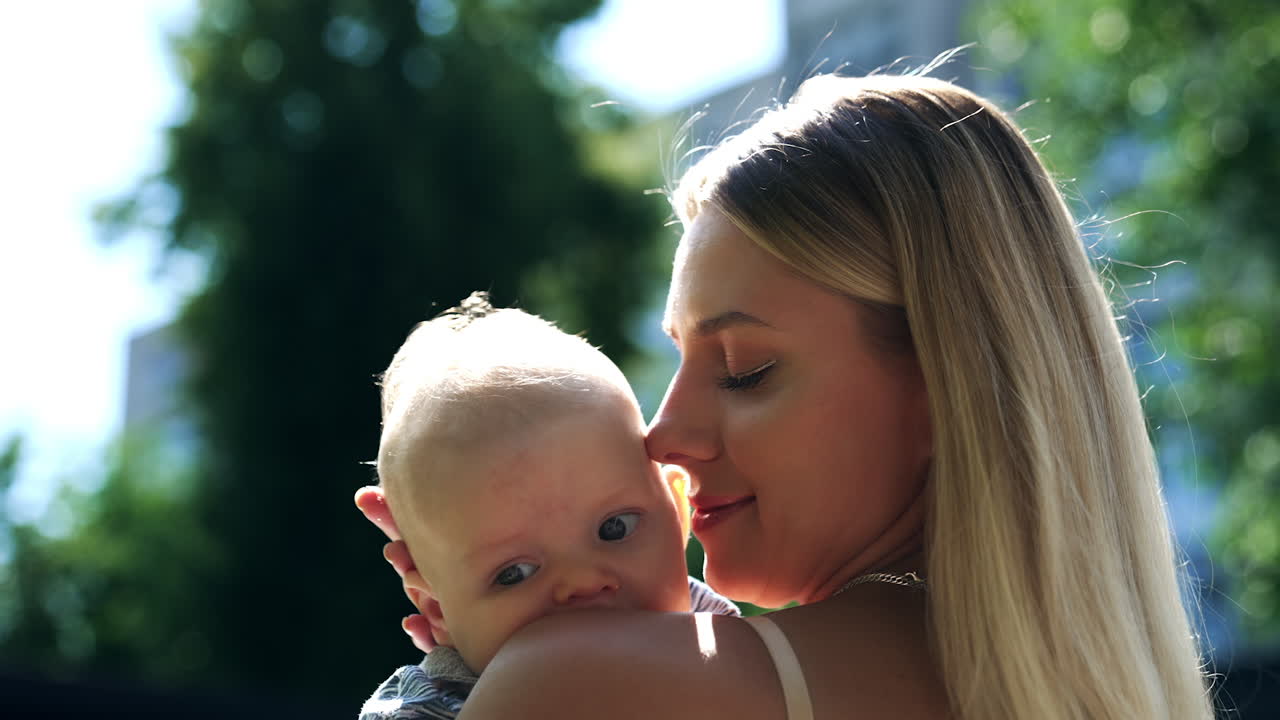 Beautiful blonde woman stands half-back to camera holding a baby. Loving mom caressing her son tenderly. Close up.