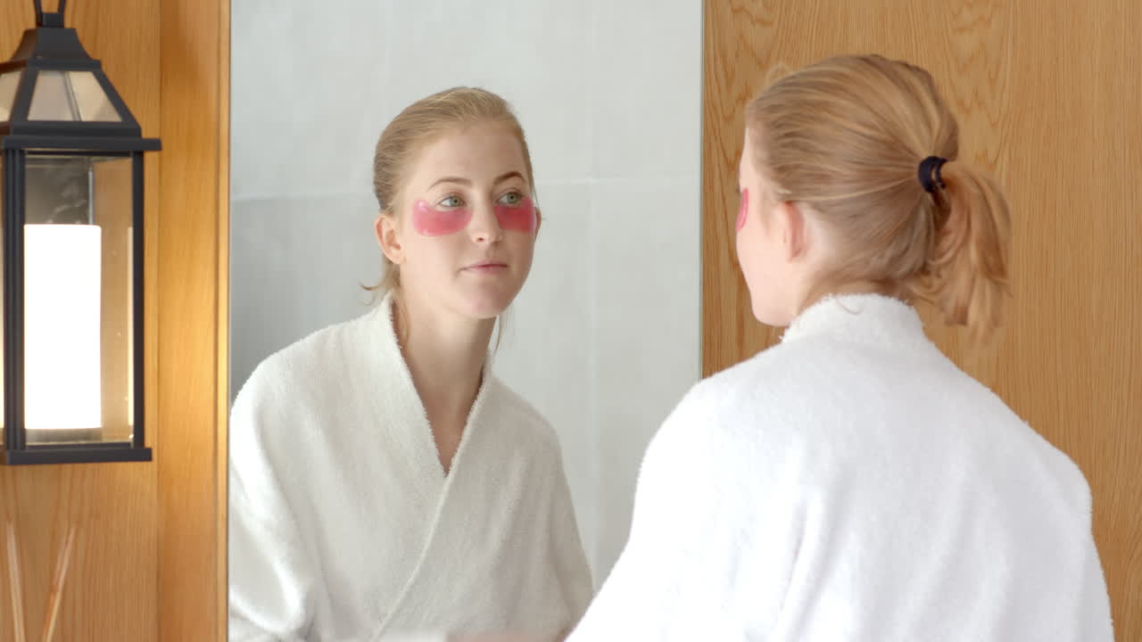 Donna in accappatoio che applica patch sotto gli occhi, guardandosi allo specchio del bagno.