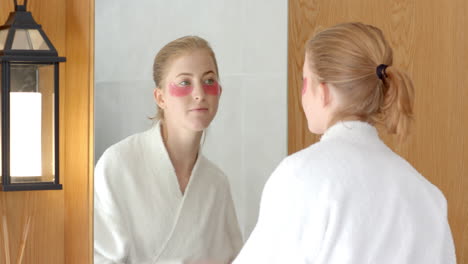 applying under-eye patches, woman in bathrobe looking in bathroom mirror