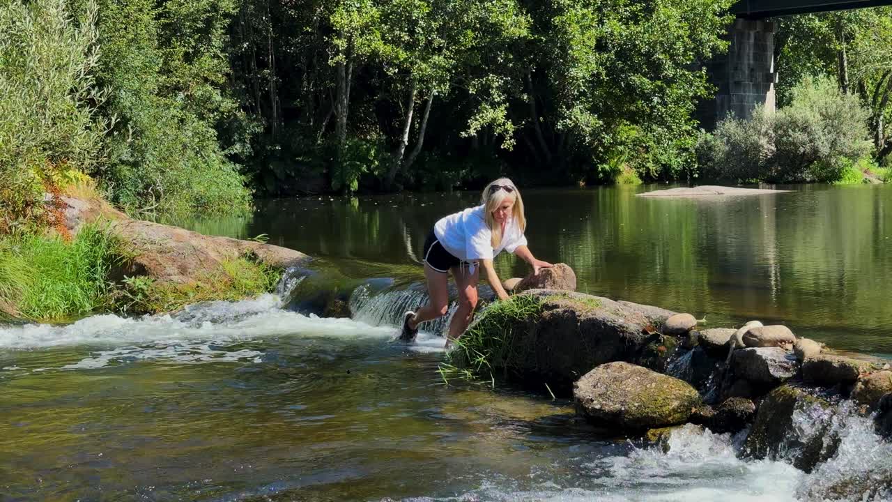 A blonde lady is trying to cross a fast flowing river over the rocks at a small waterfall