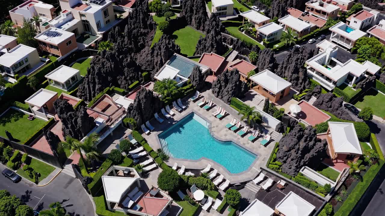 Aerial view of a luxury resort showcasing a swimming pool, sun loungers, volcanic rock formations, lush green landscaping, and surrounding residential buildings