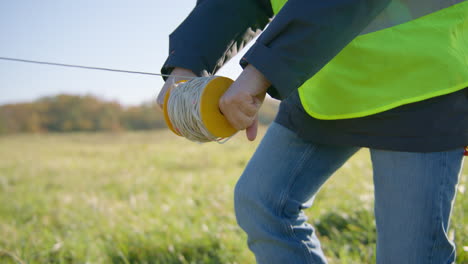 Männlicher-Ingenieur,-Der-Einen-Draht-Von-Einer-Handgehaltenen-Kabelspule-Auf-Einer-Stange-über-Das-Grasfeld-Verteilt