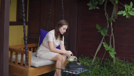 a side view of a beautiful girl sitting on a swing and having tea after pouring it from a kettle in to cup