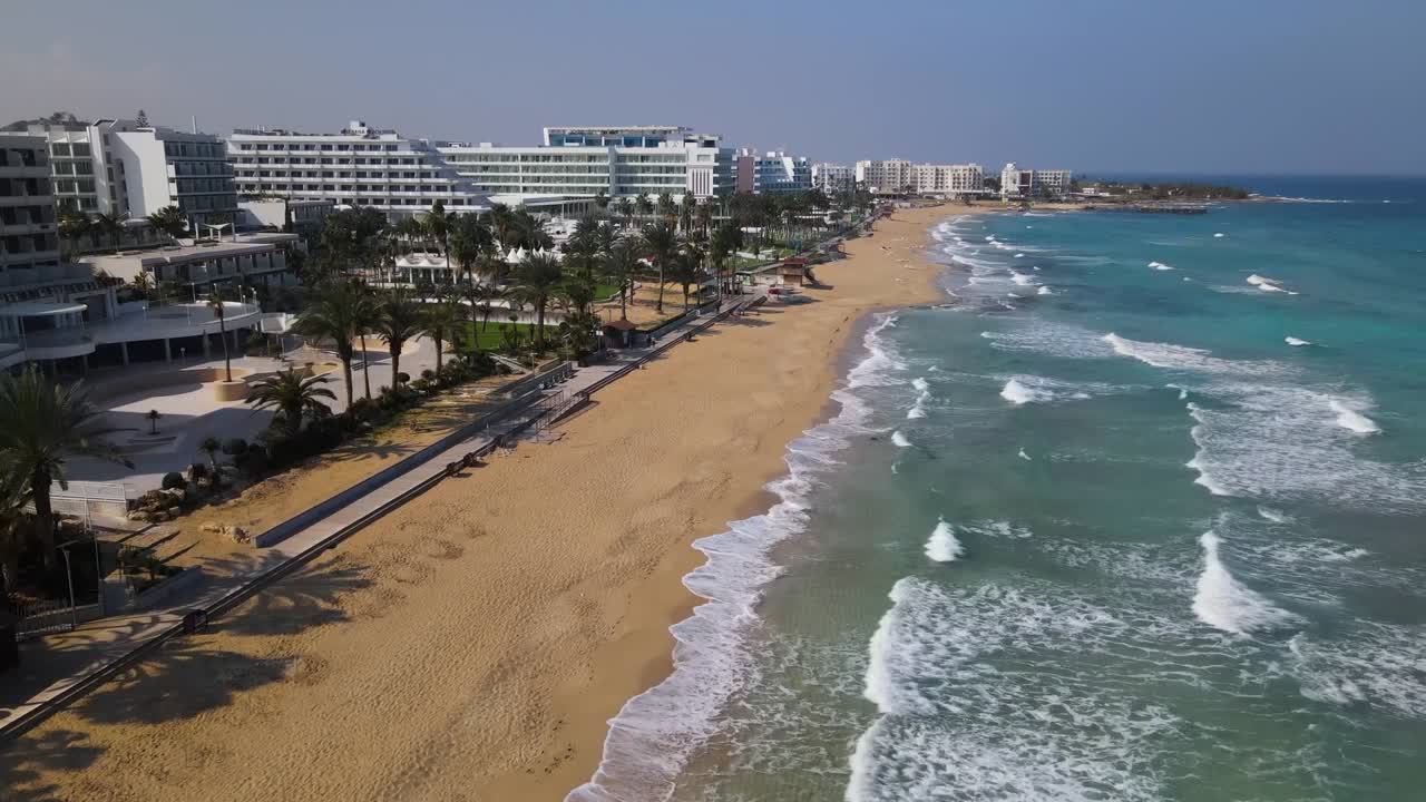 Stunning aerial footage captures Sarandë's sandy beachfront lined with palm trees, highlighting the serene promenade and resort skyline under clear blue skies and vibrant daylight