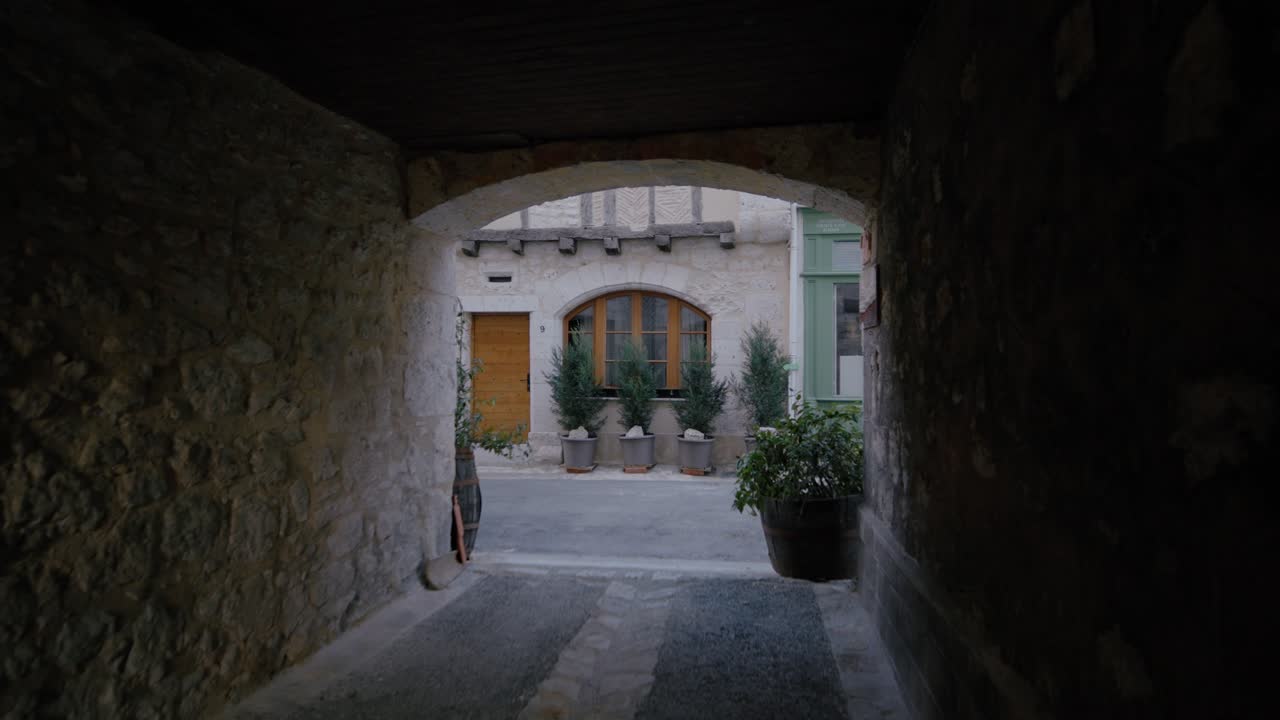 Vista attraverso un arco in pietra medievale che si affaccia su un tranquillo cortile con piante ed edifici storici a Issigeac, Dordogna. Inquadratura ampia statica