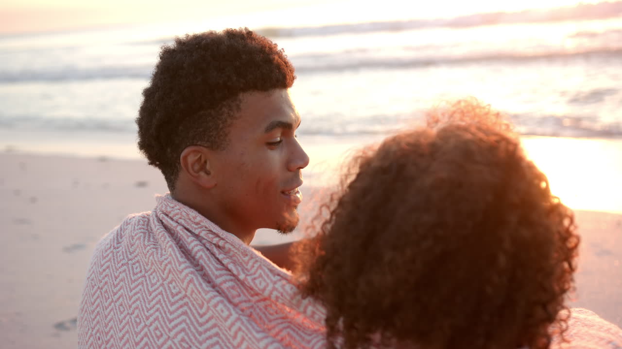 Biracial couple enjoys a serene beach sunset, wrapped in a cozy blanket