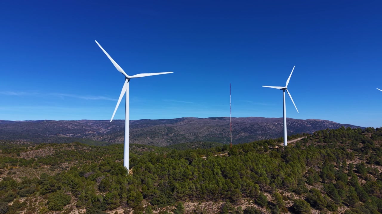 Le turbine eoliche si ergono sopra una collina boscosa sotto un cielo blu intenso con catene montuose che si estendono in lontananza, catturando energia rinnovabile in un paesaggio naturale