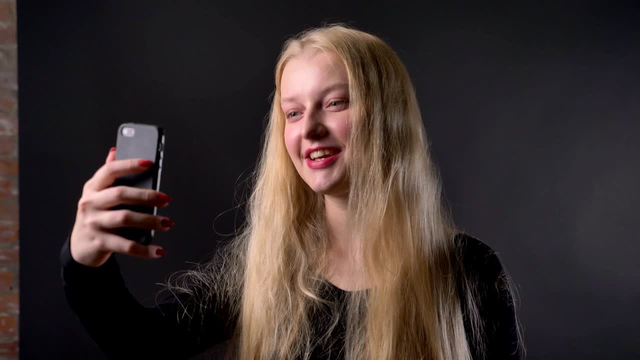 una joven rubia con lápiz labial rosa tiene una llamada de video en su teléfono inteligente, concepto de comunicación, fondo gris