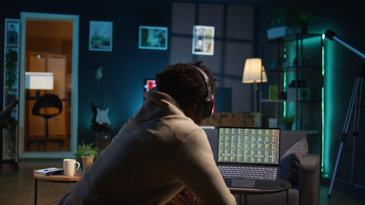 Black guy listens to music and reads from tablet on the couch, waiting for stock market trends to grow and increase his income. Young adult follows financial figures on laptop, investment. Camera B.