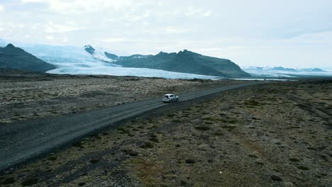 Drone-shot-circling-around-a-car-driving-through-a-scenic-fields-of-Icelandic-lava-with-the-view-of-glacier-in-the-background,-Iceland