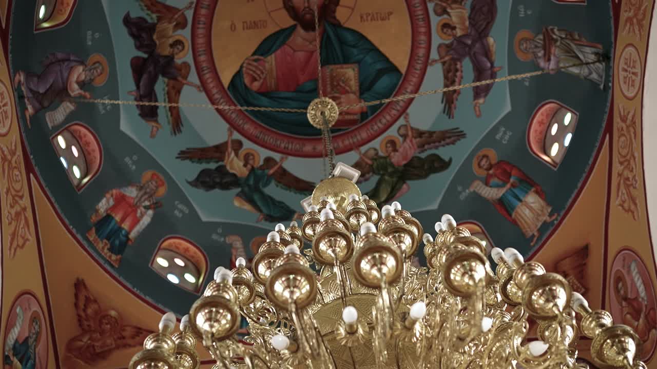 Das Innere einer Kirche zeigt eine detaillierte Decke mit religiösen Gemälden. Ein großer Kronleuchter hängt unter dem farbenprächtigen Kunstwerk. Menschen bewundern die Umgebung