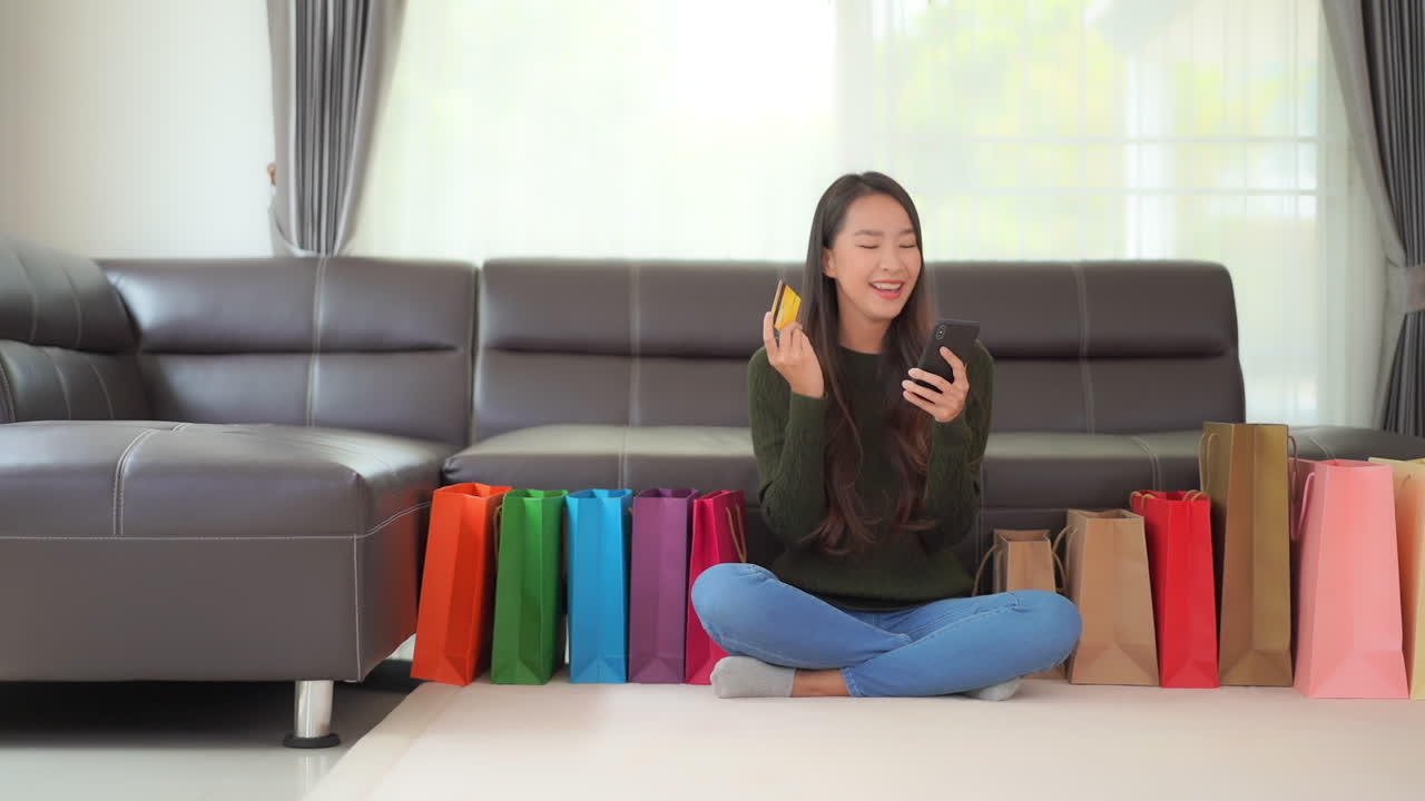 Shopping with a smartphone and credit card while at home a young, attractive Asian woman is surrounded by colorful shopping bags