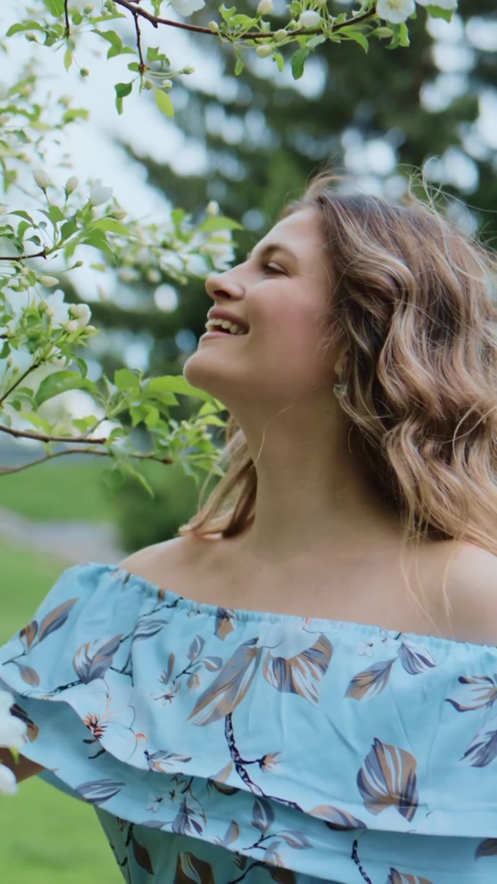 Una giovane donna sorride dolcemente all'aperto in una luce soffusa. I suoi capelli fluiscono naturalmente, gli occhi sono dolcemente chiusi. Indossa un abito blu floreale con spalle scoperte. La mano tocca i fiori bianchi in fiore.