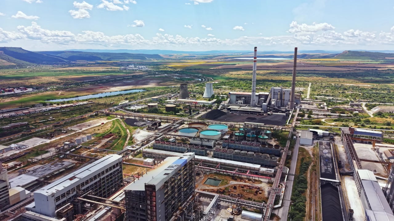 Industrial site located near a river and hills shows large structures and machinery under a bright sky with clouds during the day