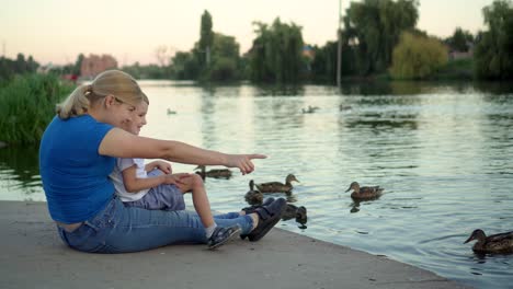 mother and child in medical masks feed ducks on the lake 01