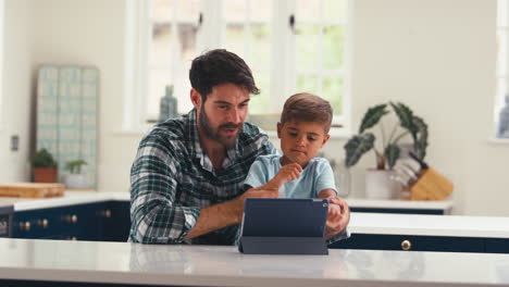 Padre-En-Casa-En-La-Cocina-Con-Su-Hijo-Transmitiendo-O-Jugando-En-Una-Tableta-Digital