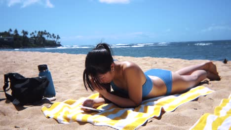 Lone-young-woman-sun-tanning-at-a-picturesque-beach-while-using-smartphone