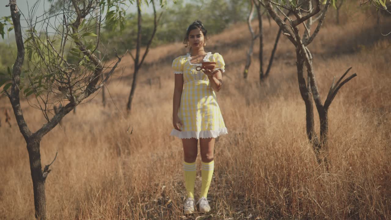 Un'inquadratura con messa a fuoco a slittamento su una donna asiatica che indossa un bel vestito giallo, mentre tiene un vaso di argilla nel palmo del braccio teso in un campo di erba secca in una giornata estiva, India