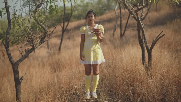 Un'inquadratura con messa a fuoco a slittamento su una donna asiatica che indossa un bel vestito giallo, mentre tiene un vaso di argilla nel palmo del braccio teso in un campo di erba secca in una giornata estiva, India