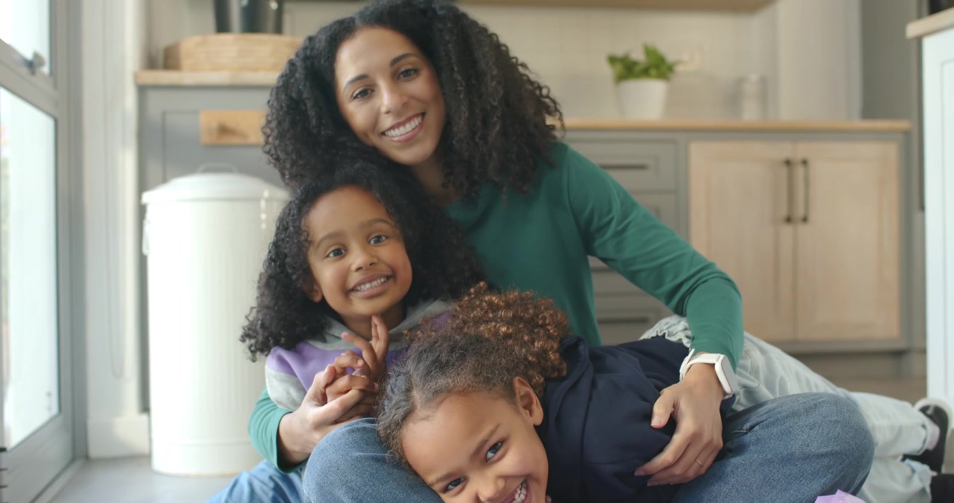 Famiglia africana di donne seduta in cucina vicino alla finestra, ridacchia e saluta la fotocamera, con un bidone trasparente visibile.