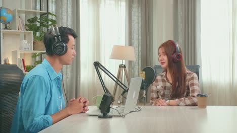 young asian woman in headphones singing into microphone while recording podcast with male host in studio