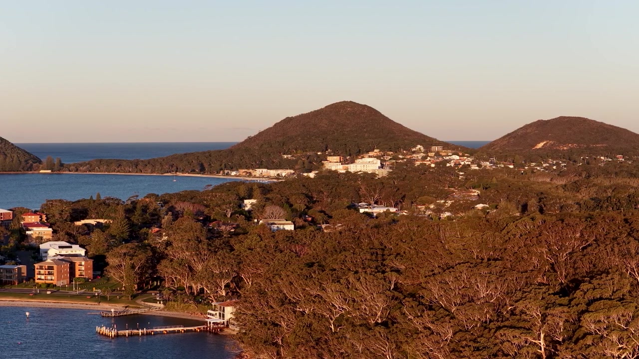 Plano aéreo panorâmico da costa de Nelson Bay, colinas arborizadas e áreas residenciais durante a hora dourada