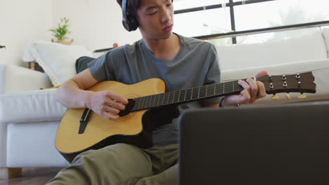 Niño-Asiático-Con-Auriculares-Tocando-La-Guitarra-Mirando-La-Computadora-Portátil-En-Casa