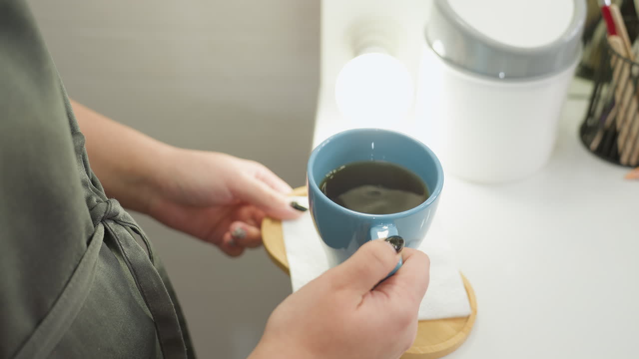 Beautician wearing apron gently holds blue ceramic tea cup on wooden tray with white napkin, showing black polished nails while carefully serving warm beverage in calm, professional environment