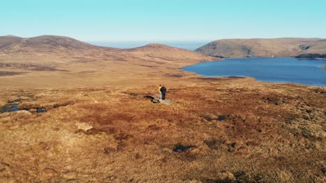 Two-hikers-on-a-rock-in-front-of-lake,-drone-rotation