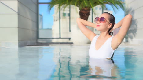 Feminine-Asian-woman-squeeze-water-out-of-hair-while-bathing-in-a-swimming-pool,-she-is-in-water-up-to-the-chest-and-touching-her-wet-hair,-face-close-up,-a-luxury-resort-in-Thailand