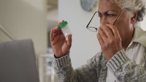 mujer mayor afroamericana sosteniendo un contenedor de medicamentos haciendo una videollamada en una computadora portátil en casa