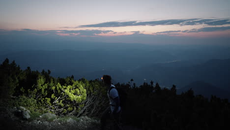 Wanderer,-Der-Mit-Wanderstöcken-Und-Stirnlampe-An-Der-Kamera-Vorbeigeht-Und-Auf-Das-Tal-Und-Die-Richtung-Blickt,-In-Der-Die-Sonne-Aufgehen-Wird