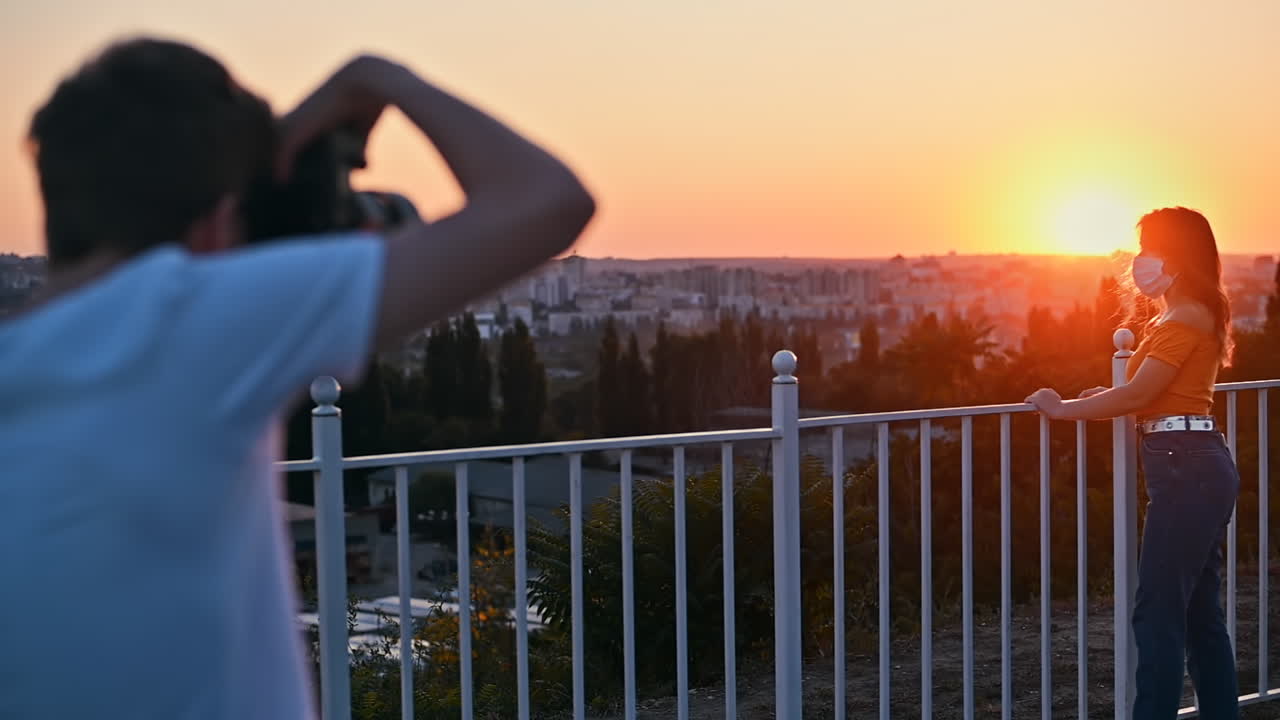 Photographer taking photos of a young woman at observation deck with city and orange sunset on background. Model wearing protective medical mask. Rear view. Corona virus idea