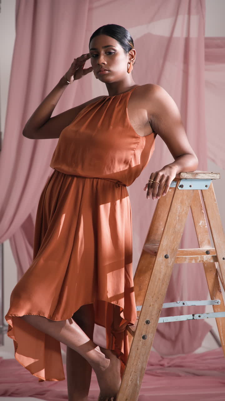 Vertical video: Model in elegant orange dress posing confidently on wooden ladder in studio